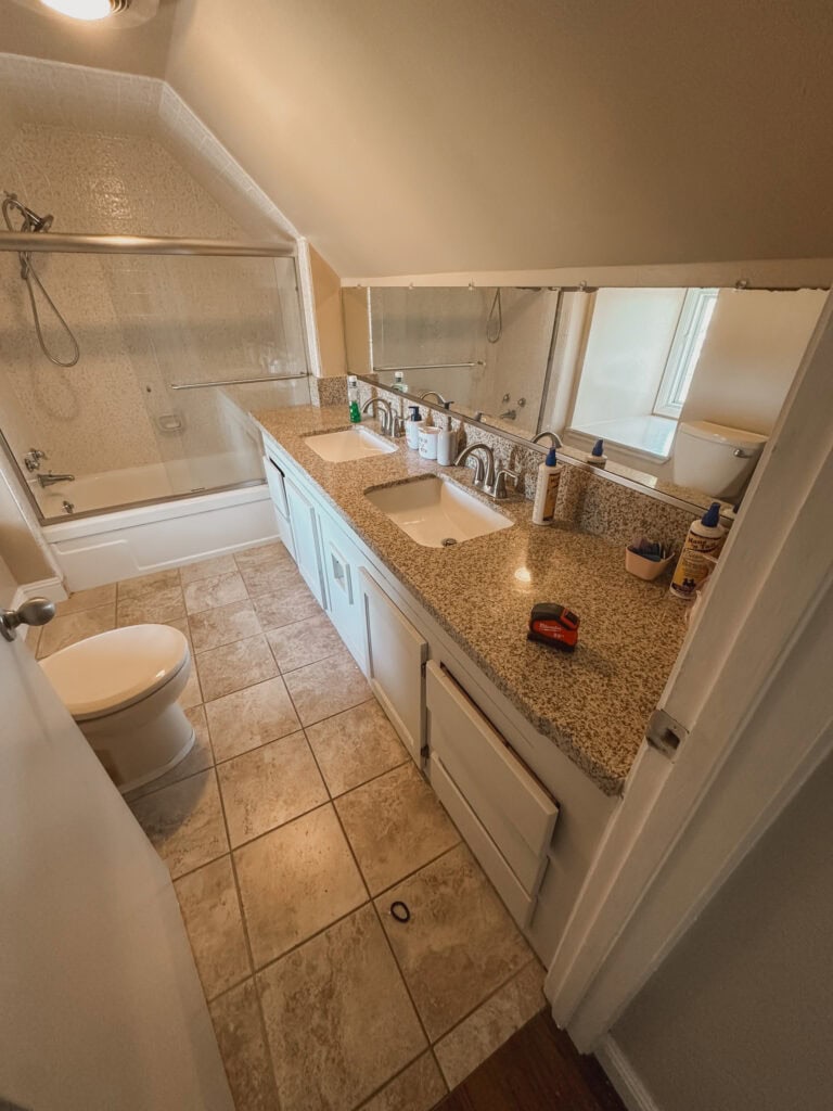 This is the before photo of my step kid's bathroom before I did a makeover. It is a small dated bathroom with slanted ceilings, brown walls, brown floor, cream tile, a glass shower door, gray vanity with brown granite countertops with a an ugly damaged mirror above.