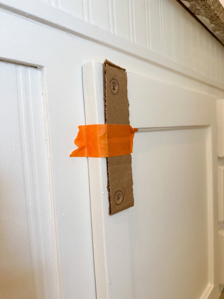 Photo of cardboard template taped with orange tape to a vanity to get pilot hole placement right while installing hardware on a bathroom vanity.